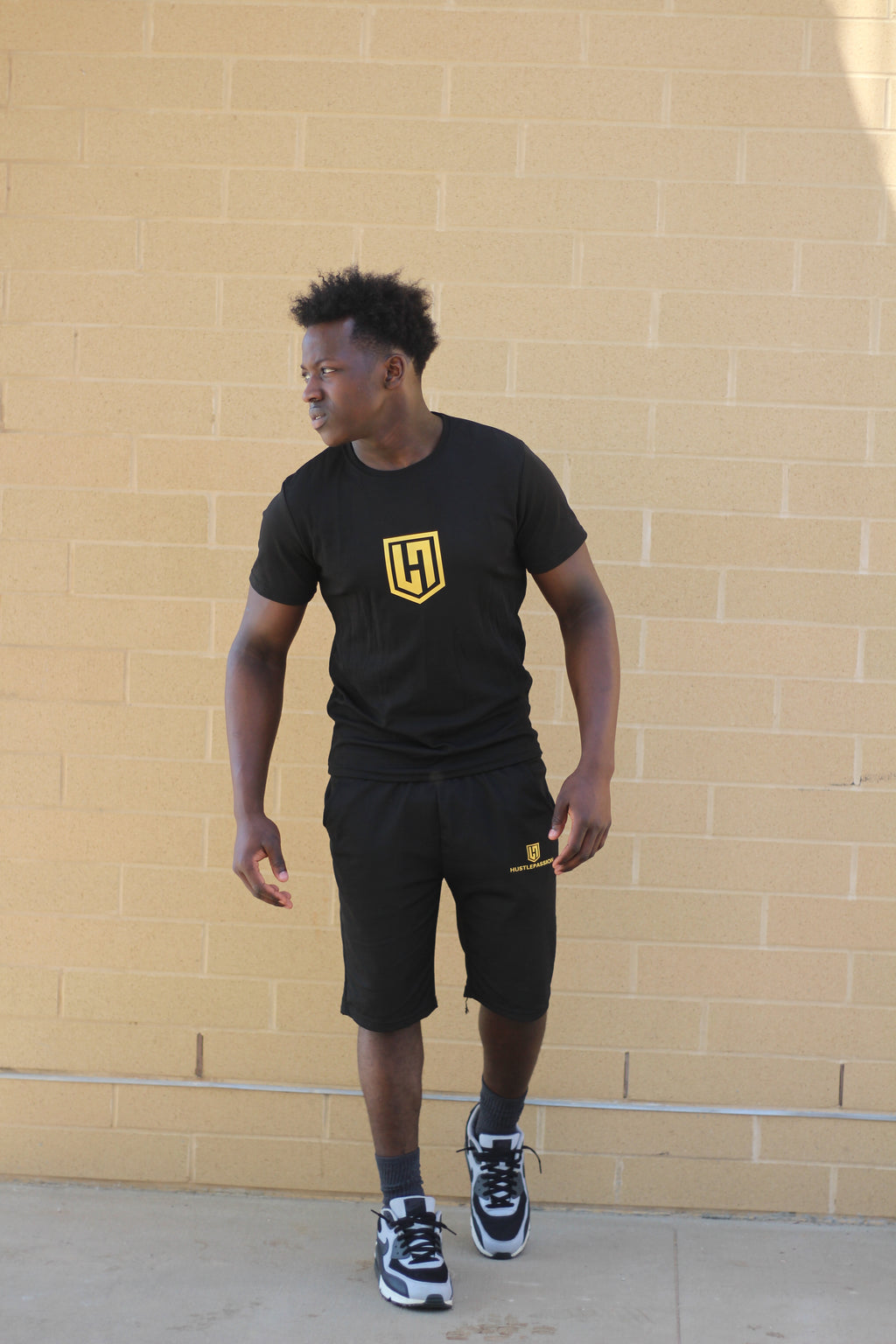 Model wearing the Hustle Passion T-shirt and shorts set in black, walking by a brick wall