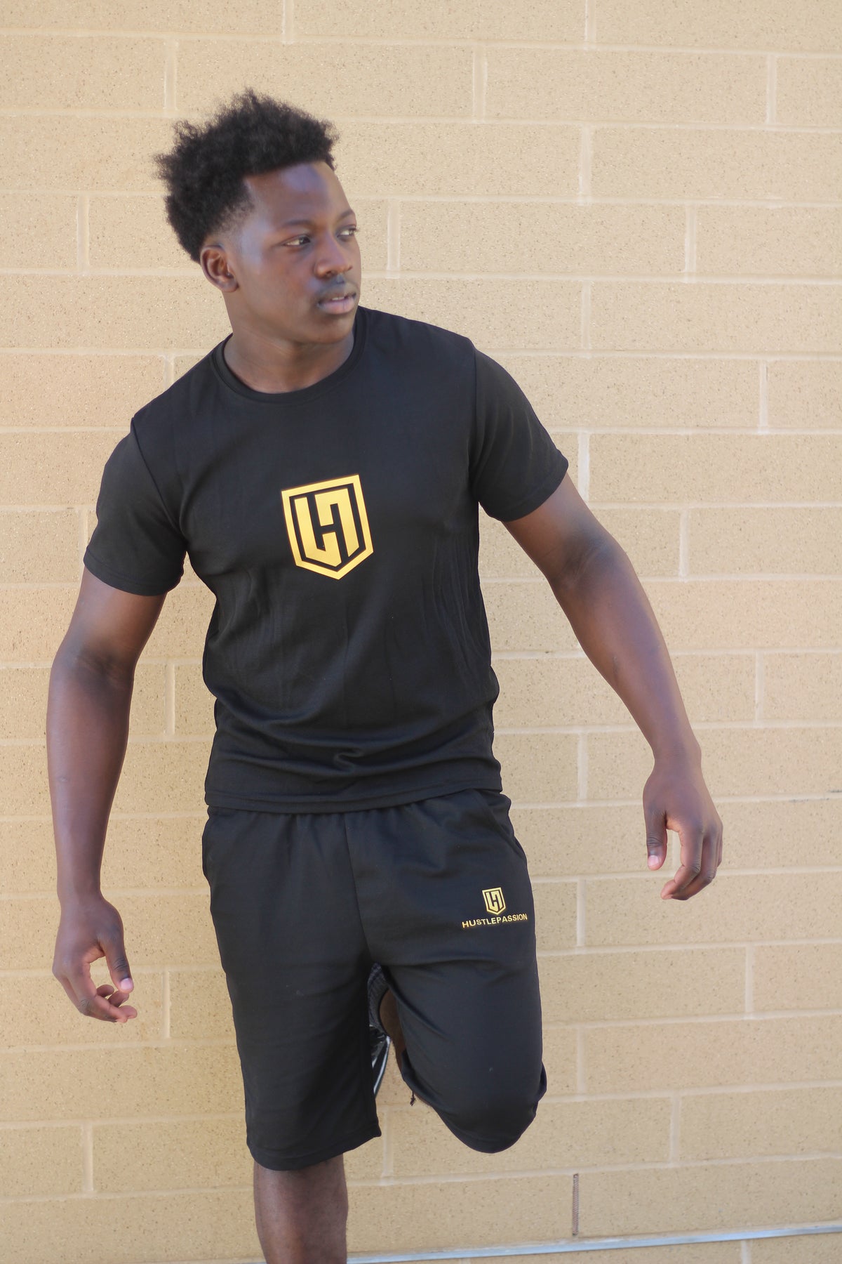 Model wearing the black Hustle Passion T-shirt and shorts set, mid-stride by a brick wall