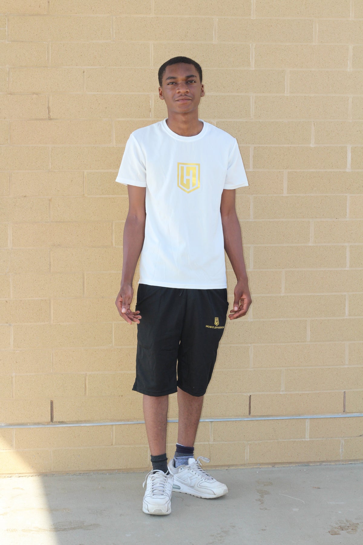 Model wearing a white Hustle Passion T-shirt with black athletic shorts, standing by a brick wall