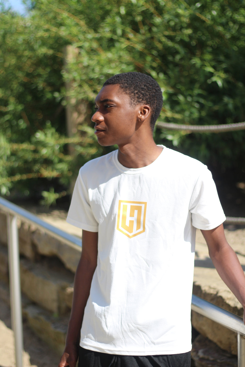 Model wearing a white Hustle Passion T-shirt with gold logo, standing outdoors among trees