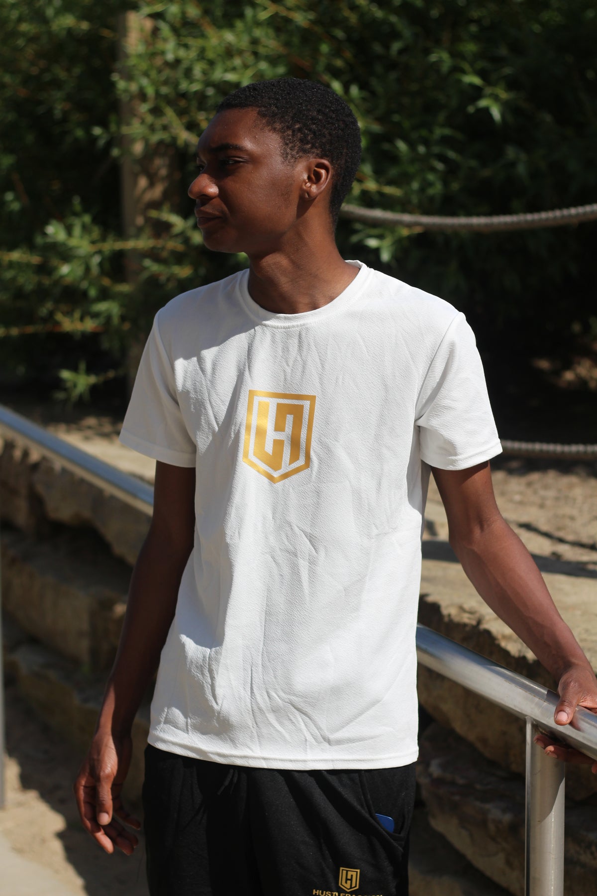 Model in a white Hustle Passion T-shirt and black shorts standing by an outdoor railing