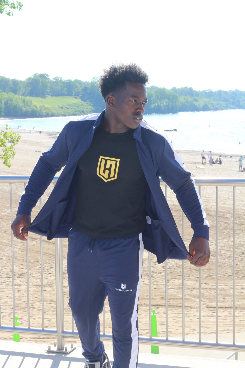 Model by the lake wearing the navy blue tracksuit jacket open over a black Hustle Passion T-shirt