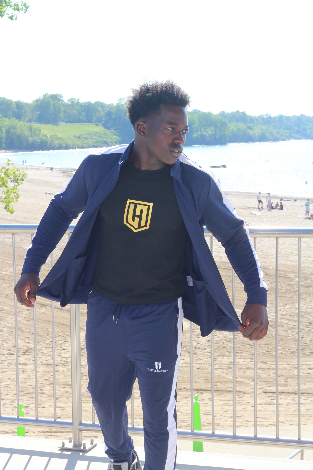 Model by the lake wearing the navy blue tracksuit jacket open over a black Hustle Passion T-shirt