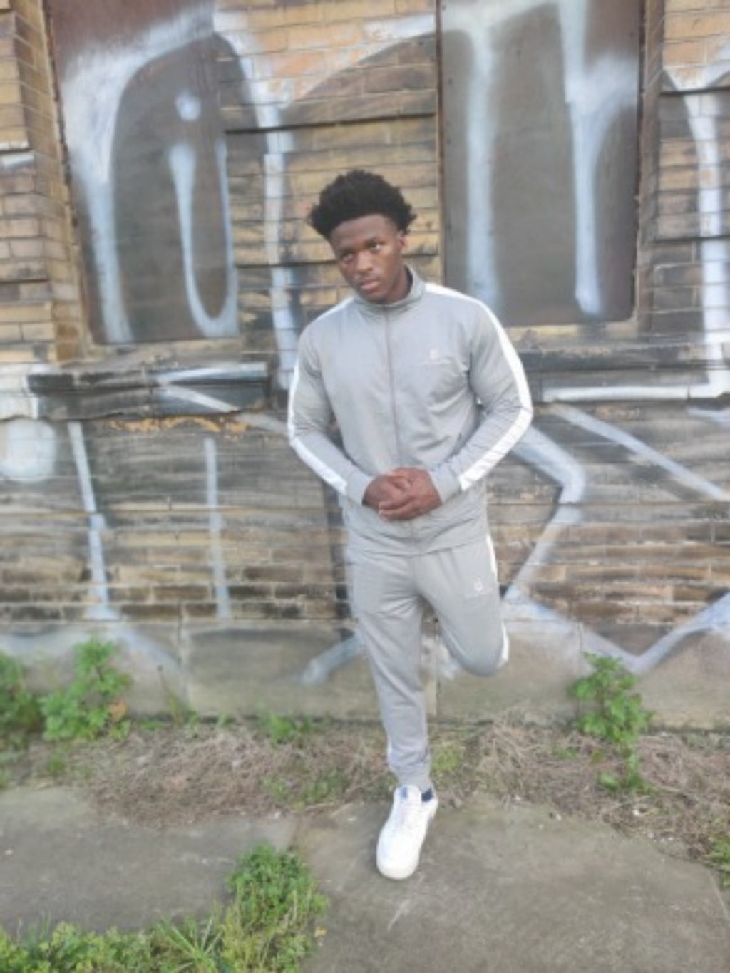 Model in the light grey tracksuit leaning against a graffiti wall, one foot raised