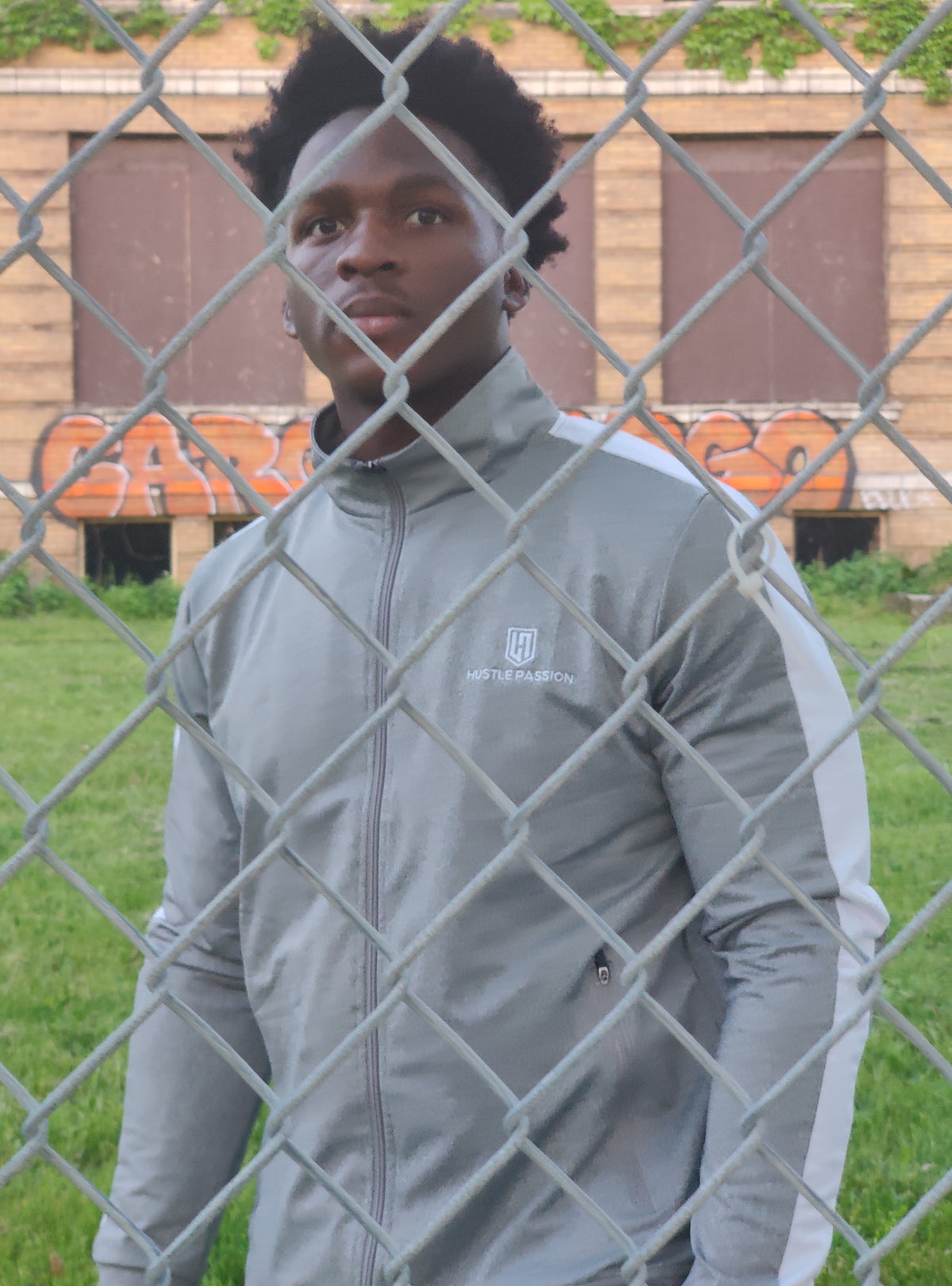 Close-up of a model wearing the light grey tracksuit jacket behind a chain-link fence