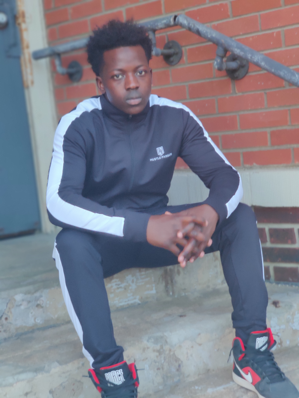 Model sitting on steps wearing the Hustle Passion pitch black tracksuit with white side stripes